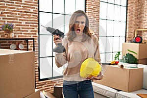 Young woman holding drill at new home in shock face, looking skeptical and sarcastic, surprised with open mouth