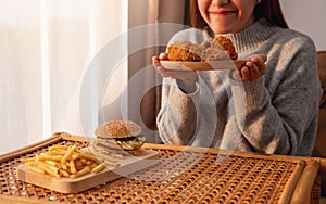 A young woman holding and eating fried chicken with hamburger and french fries on the table at home