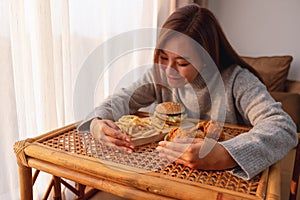 A young woman holding and eating a hamburger, french fries and fried chicken on the table at home