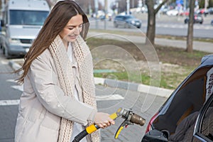 Young woman holding a fuel nozzle in her hand while refueling car at gas station.