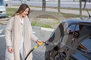 Young woman holding a fuel nozzle in her hand while refueling car at gas station.