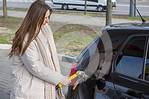 Young woman holding a fuel nozzle in her hand while refueling car at gas station.