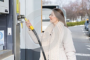 Young woman holding a fuel nozzle in her hand while refueling car at gas station.