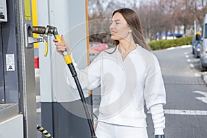 Young woman holding a fuel nozzle in her hand while refueling car at gas station.