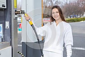 Young woman holding a fuel nozzle in her hand while refueling car at gas station.