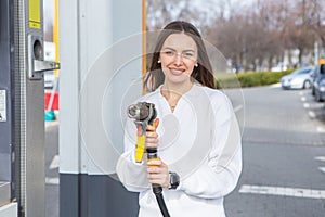 Young woman holding a fuel nozzle in her hand while refueling car at gas station.
