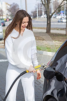 Young woman holding a fuel nozzle in her hand while refueling car at gas station.