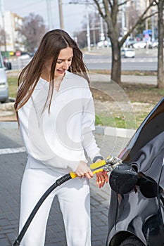 Young woman holding a fuel nozzle in her hand while refueling car at gas station.