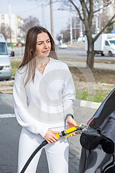 Young woman holding a fuel nozzle in her hand while refueling car at gas station.
