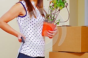 Young woman holding green plant and grabbing suitcase handle with the other, boxes stacked background, moving in concept