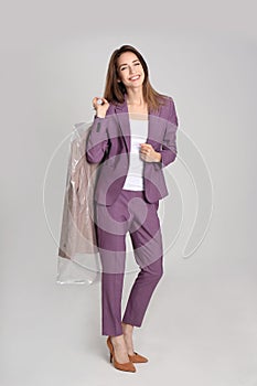 Young woman holding hanger with jacket in plastic bag on grey background. Dry-cleaning service