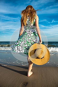 Young woman holding hat and walking on beach sand to the ocean