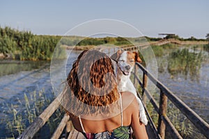 Young woman holding her cute small jack russell terrier on shoulder. Standing in a wood pier in a natural park. Tourism, travel