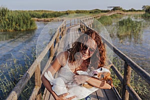 Young woman holding her cute small jack russell terrier on shoulder. Standing in a wood pier in a natural park. Tourism, travel