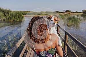 Young woman holding her cute small jack russell terrier on shoulder. Standing in a wood pier in a natural park. Tourism, travel