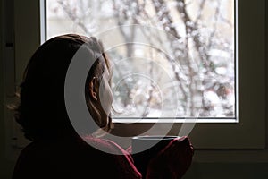 Young woman holding a hot coffee or tea cup looking out of the window in a snowy winter day