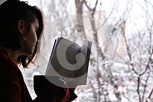 Young woman holding a hot coffee or tea cup, reading a book by the window in a snowy winter day