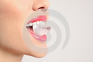 Young woman holding ice cube in mouth on light background, closeup
