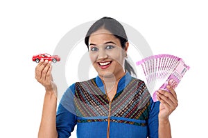 Young woman holding indian currency and toy car against white