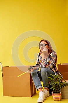 Young woman holding a measuring tape sitting on a tool box, between boxes for moving