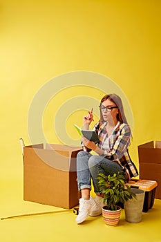 Young woman holding a notepad sitting on a tool box, between boxes for moving