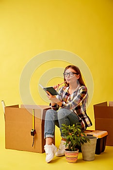 Young woman holding a notepad sitting on a tool box, between boxes for moving