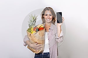 Young woman holding paper bag full of groceries and showing her mobile phone, Order online concept. shopping app