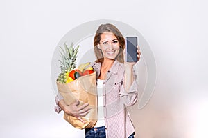 Young woman holding paper bag full of groceries and showing her mobile phone, Order online concept. shopping app