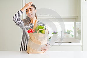 Young woman holding paper bag full of groceries stressed with hand on head, shocked with shame and surprise face, angry and