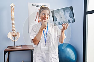 Young woman holding pelvis radiography serious face thinking about question with hand on chin, thoughtful about confusing idea