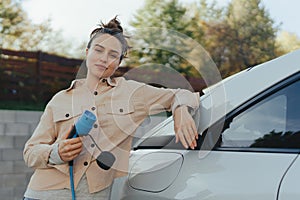 Young woman holding power supply cable from her electric car, prepared for charging it in home, sustainable and economic