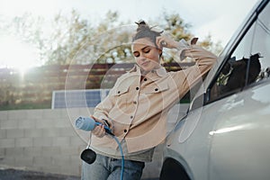 Young woman holding power supply cable from her electric car, prepared for charging it in home, sustainable and economic