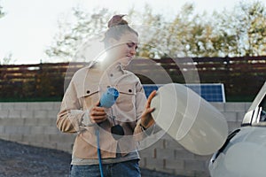 Young woman holding power supply cable from her electric car, prepared for charging it in home, sustainable and economic