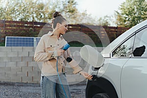 Young woman holding power supply cable from her electric car, prepared for charging it in home, sustainable and economic