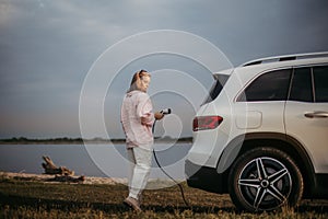 Young woman holding power supply cable from her electric car, prepared for charging it sustainable and economic