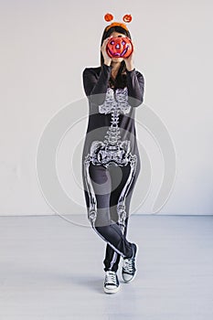 young woman holding a pumpkin and smiling. Wearing a black and white skeleton costume. Halloween concept. Indoors. Lifestyle