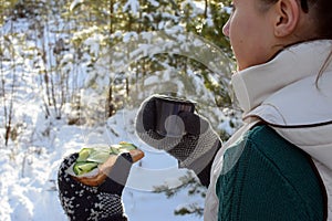 Young woman holding sandwitch and coffee mug in winter forest.