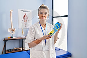 Young woman holding shoe insole at physiotherapy clinic relaxed with serious expression on face