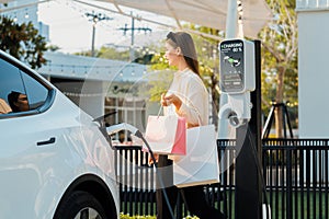 Young woman holding shopping bag recharge EV car. Expedient