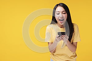 Young woman holding smartphone and her hand with shocked amazed for success or get good news over isolated yellow