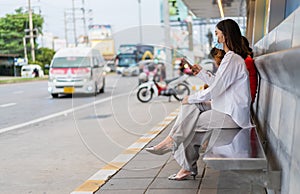 Young woman holding smartphone and waiting for bus at bus stop in city street and wearing face mask protective for spreading of