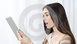 Young woman holding a tablet and looking at it surprised with her mouth open on white background