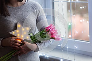 Young woman holding tender spring bouquet of pink tulips and glowing LED star near window with magic bokeh lights