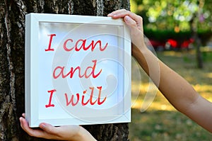 Young woman holding by a tree , a framed  motivational message stating I can and I will
