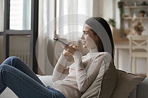 Young woman holds cellphone talks on speakerphone relaxing on sofa