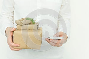 Young woman holds in hands stacked Christmas gift boxes in craft paper with juniper and smartphone. Holiday corporate presents