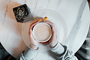 Young woman holds in her hands a cup with a hot sweet cappuccino sitting at a table in a cafe. Great morning for coffee gourmets.