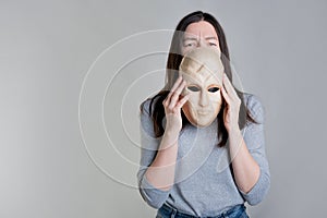 A young woman holds a mask in her hands, hiding her face half, an exhausted look over the mask.