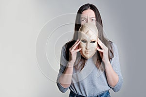 A young woman holds a mask in her hands, hiding her face half, a frightened look over the mask.