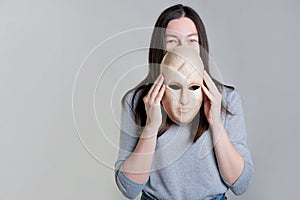 A young woman holds a mask in her hands, hiding her face half, a sly look over the mask.